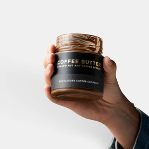 Hand holding a glass jar of Bootlegger Coffee Coffee Butter, a cashew nut and coffee spread with a black label.